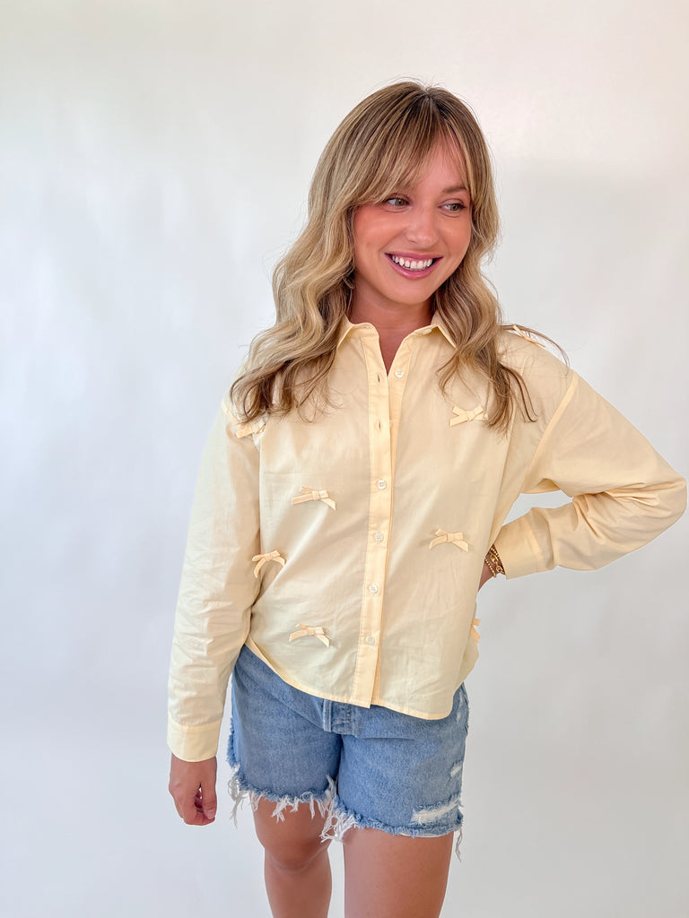Model wearing a pale yellow blouse with small bow details on the front and shoulder seams