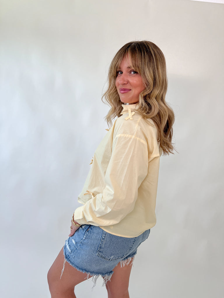 Model wearing a pale yellow blouse with small bow details on the front and shoulder seams