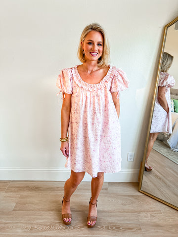 Light pink floral babydoll dress with ruffle neckline, short puff sleeves with tie details, and a loose, flowy silhouette. Perfect for a feminine and effortless boutique look