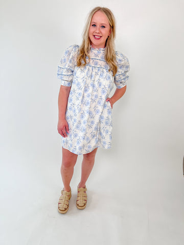 Woman wearing a mini dress with blue floral print, puff sleeves, and high ruffle neckline