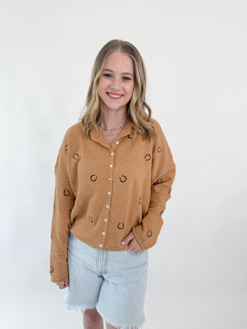 Woman wearing a caramel knit cardigan with embroidered horseshoes and button front.