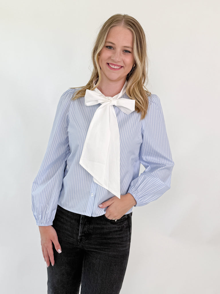 Woman wearing a blue and white pinstripe blouse with a large white bow and puff sleeves