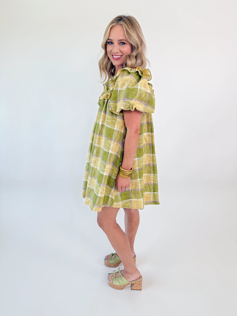 Model wearing the Meadow Plaid Ruffle Mini Dress in green, yellow, and lavender plaid with ruffle neckline and short puff sleeves