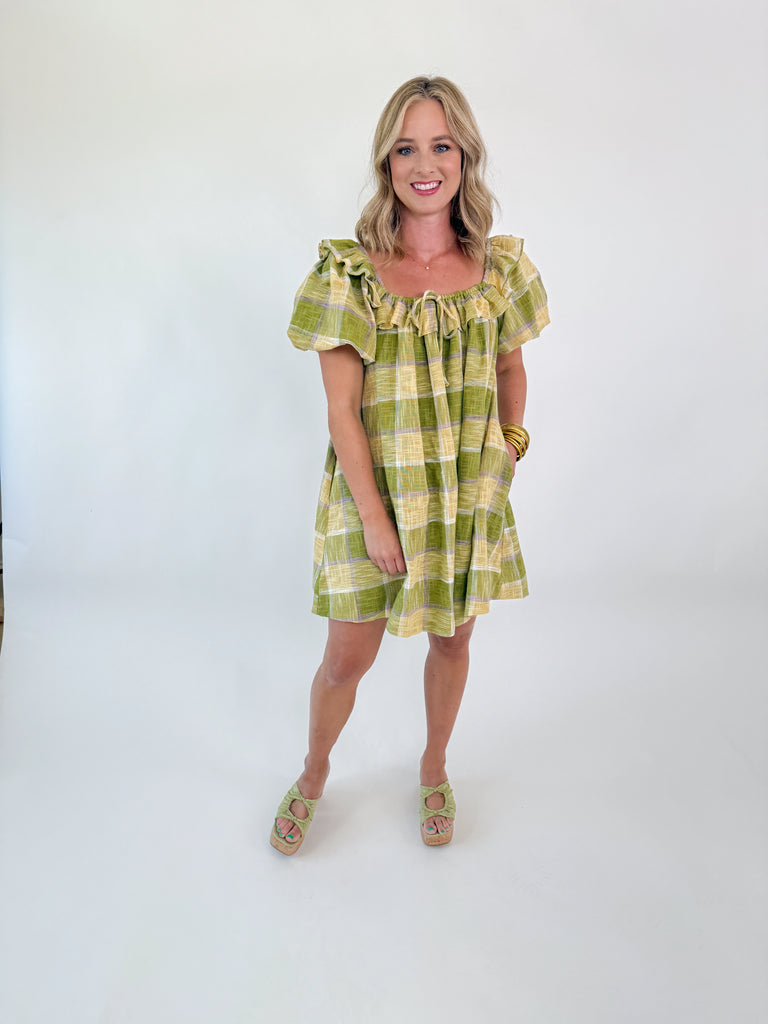 Model wearing the Meadow Plaid Ruffle Mini Dress in green, yellow, and lavender plaid with ruffle neckline and short puff sleeves