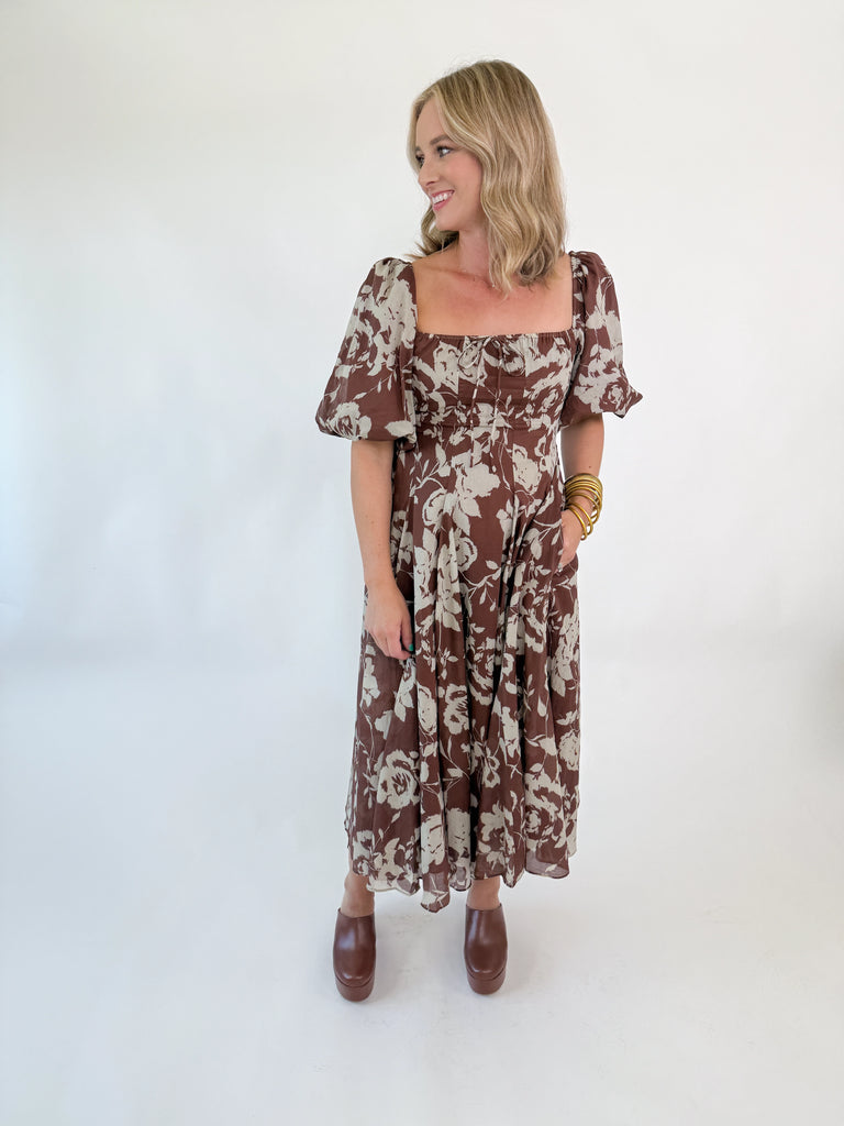 Woman wearing mocha brown puff-sleeve midi dress with cream floral print, square neckline, fitted bodice, and flowy skirt