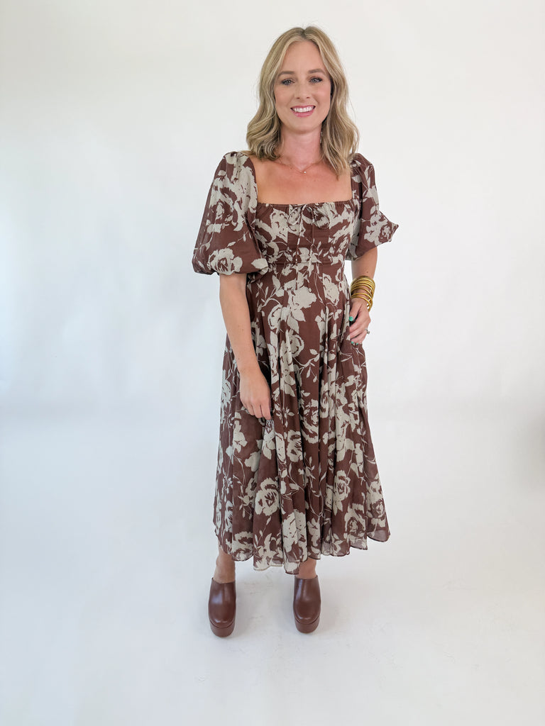 Woman wearing mocha brown puff-sleeve midi dress with cream floral print, square neckline, fitted bodice, and flowy skirt