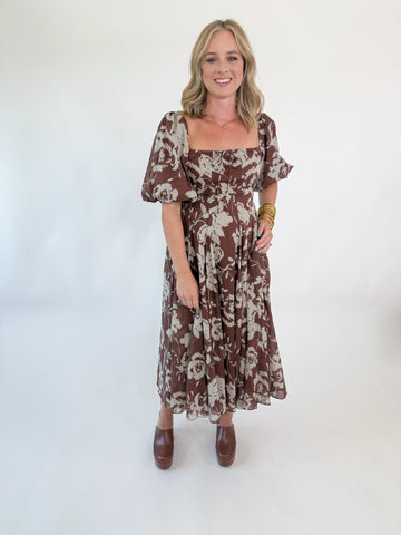 Woman wearing mocha brown puff-sleeve midi dress with cream floral print, square neckline, fitted bodice, and flowy skirt