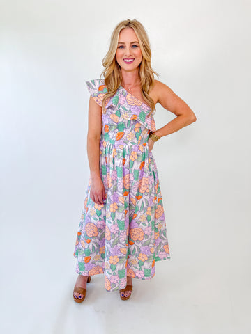 Woman wearing a pastel floral one-shoulder maxi dress with ruffle detail and a smocked waist