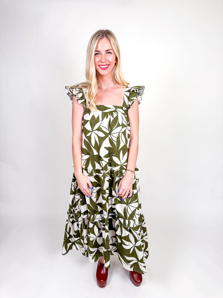 Woman wearing an olive and ivory leaf-print maxi dress with ruffle straps, square neckline, tiered skirt, and open back bow