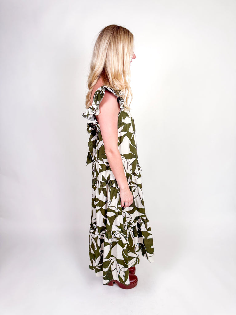 Woman wearing an olive and ivory leaf-print maxi dress with ruffle straps, square neckline, tiered skirt, and open back bow