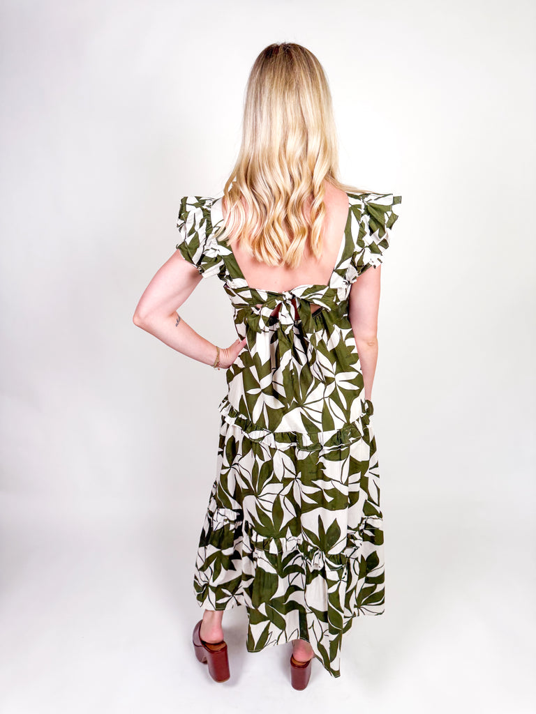 Woman wearing an olive and ivory leaf-print maxi dress with ruffle straps, square neckline, tiered skirt, and open back bow