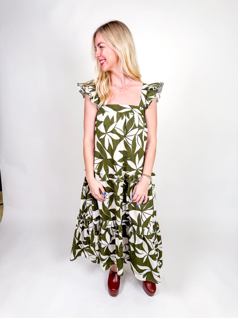 Woman wearing an olive and ivory leaf-print maxi dress with ruffle straps, square neckline, tiered skirt, and open back bow
