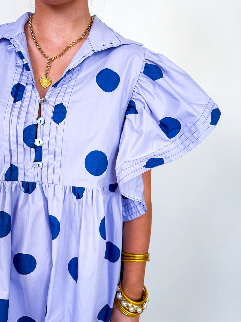 Woman wearing the Polka Daydream Dress, a periwinkle mini with navy polka dots, flutter sleeves, collared neckline, and tiered skirt