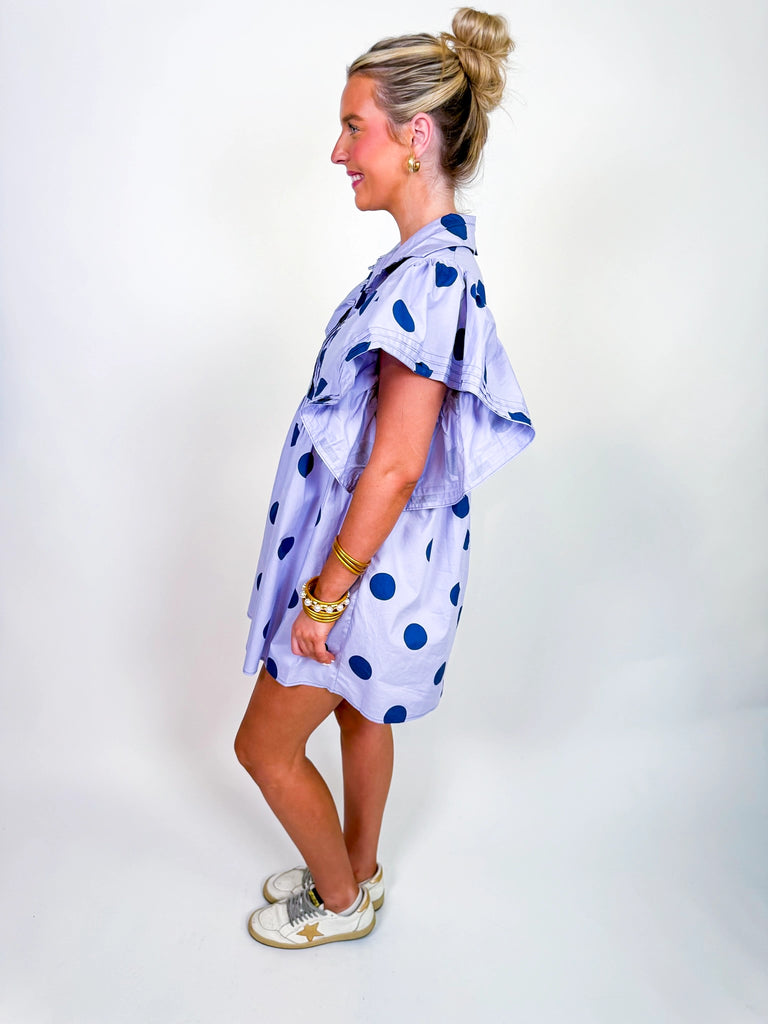 Woman wearing the Polka Daydream Dress, a periwinkle mini with navy polka dots, flutter sleeves, collared neckline, and tiered skirt
