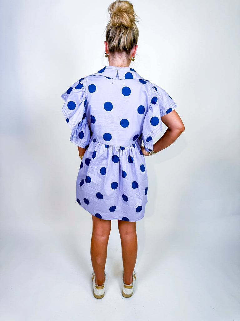 Woman wearing the Polka Daydream Dress, a periwinkle mini with navy polka dots, flutter sleeves, collared neckline, and tiered skirt