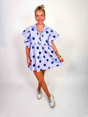 Woman wearing the Polka Daydream Dress, a periwinkle mini with navy polka dots, flutter sleeves, collared neckline, and tiered skirt