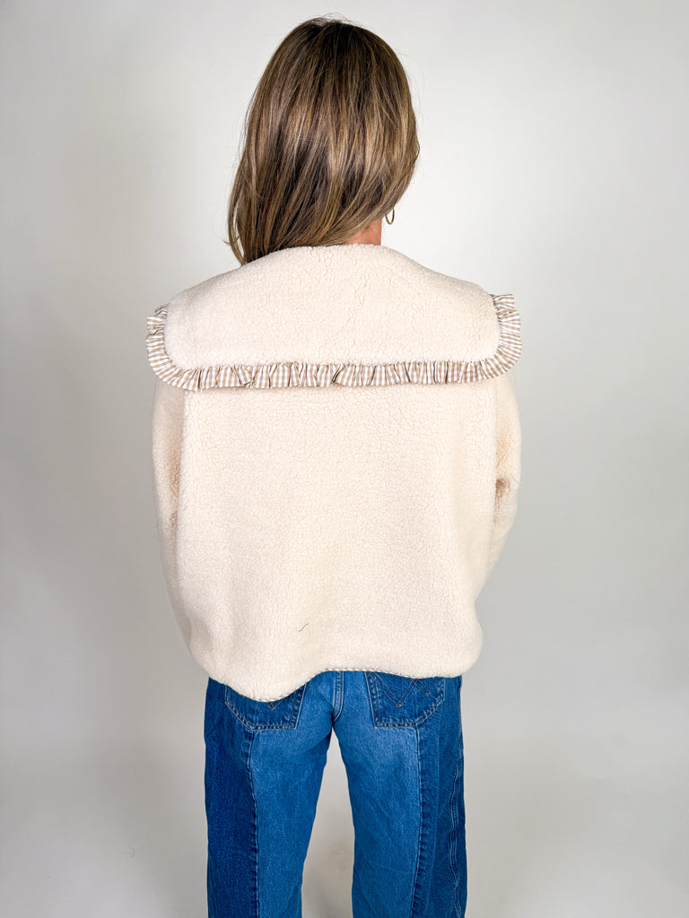 Woman wearing a cozy sherpa jacket with oversized ruffle-trimmed collar, contrast striped detail, and button front closure in blue, green, and latte.