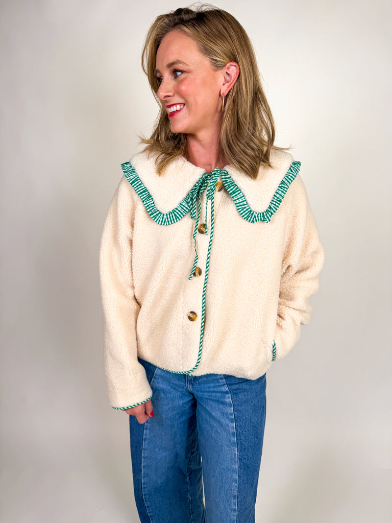 Woman wearing a cozy sherpa jacket with oversized ruffle-trimmed collar, contrast striped detail, and button front closure in blue, green, and latte.