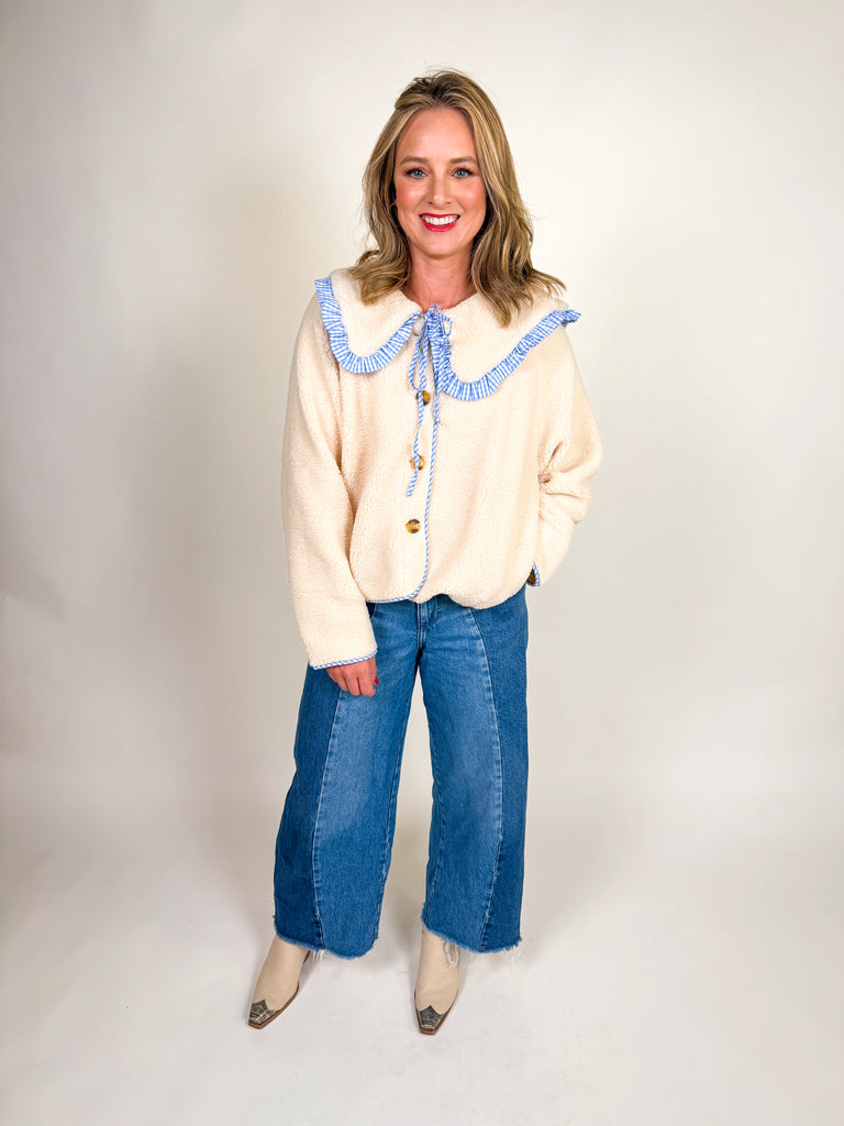 Woman wearing a cozy sherpa jacket with oversized ruffle-trimmed collar, contrast striped detail, and button front closure in blue, green, and latte.