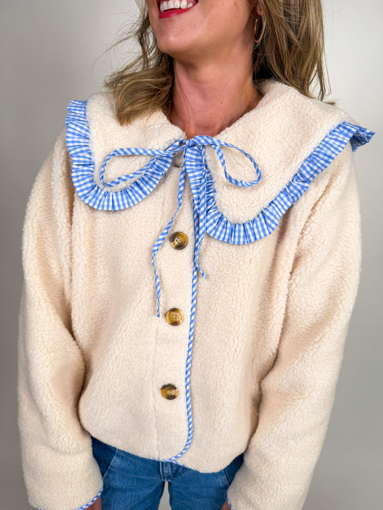 Woman wearing a cozy sherpa jacket with oversized ruffle-trimmed collar, contrast striped detail, and button front closure in blue, green, and latte.