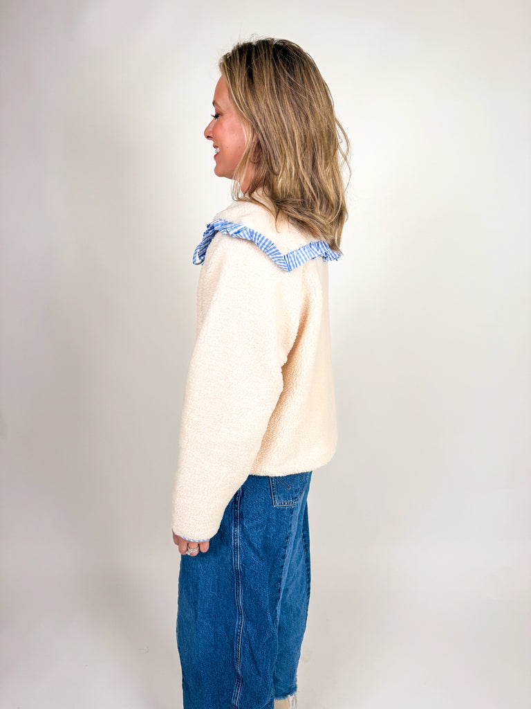 Woman wearing a cozy sherpa jacket with oversized ruffle-trimmed collar, contrast striped detail, and button front closure in blue, green, and latte.