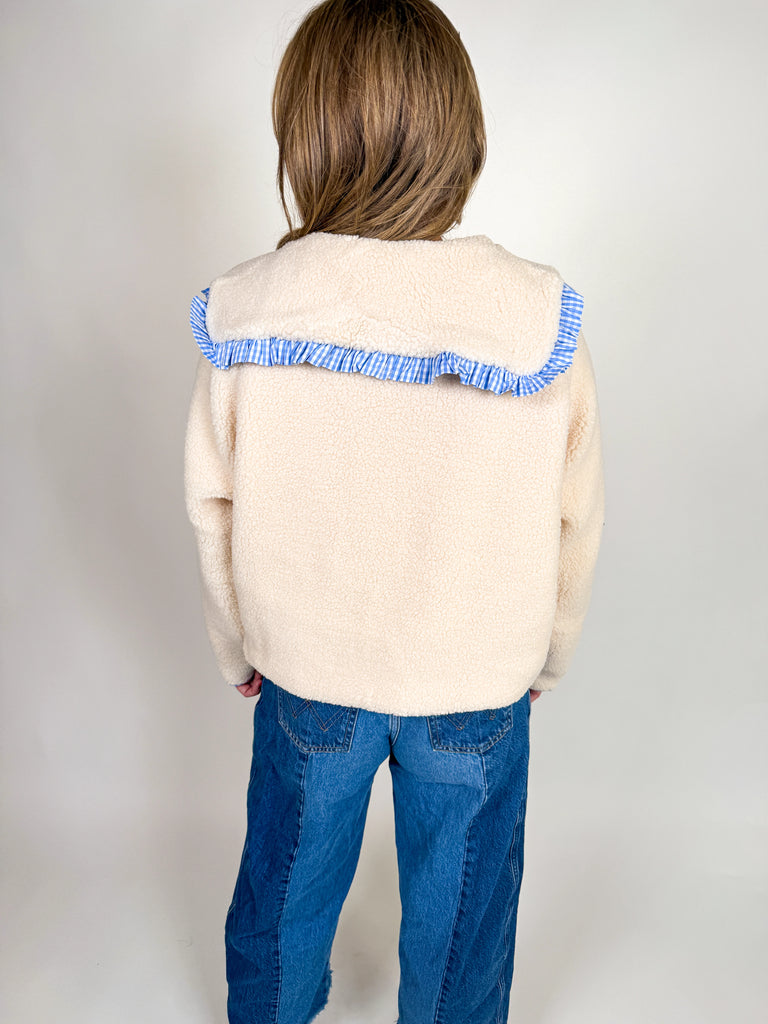 Woman wearing a cozy sherpa jacket with oversized ruffle-trimmed collar, contrast striped detail, and button front closure in blue, green, and latte.