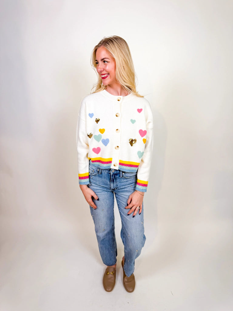 Woman wearing the Sweetheart Charm Cardigan, a white knit sweater with colorful heart appliqués and gold sequin hearts, finished with striped cuffs and hem.