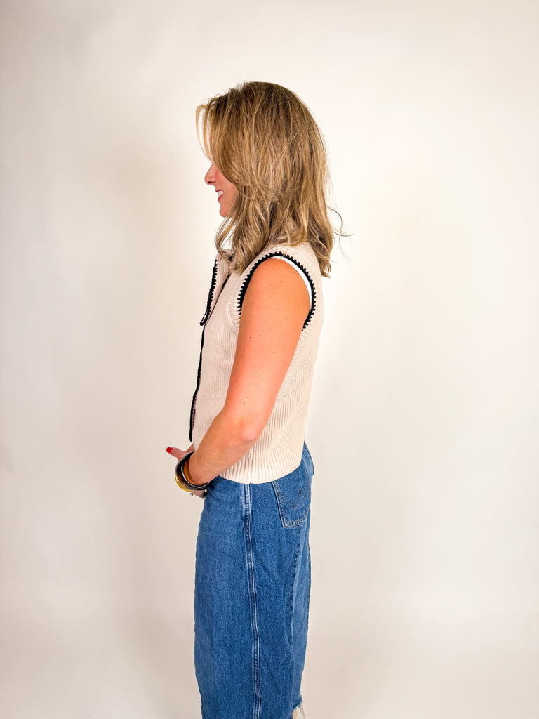 Woman wearing a cream ribbed knit vest with black contrast trim, button front, and bow neckline detail