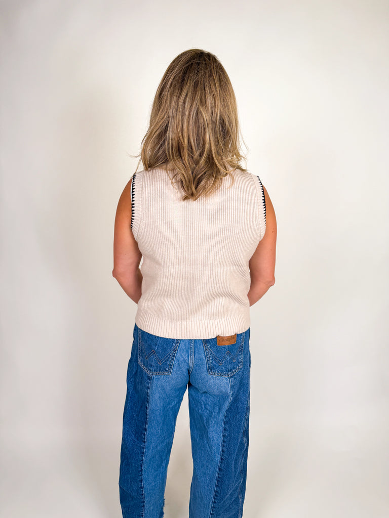 Woman wearing a cream ribbed knit vest with black contrast trim, button front, and bow neckline detail