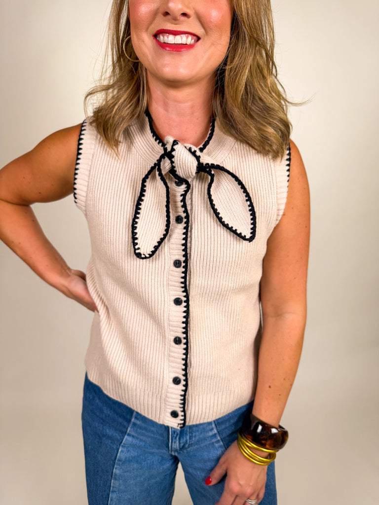 Woman wearing a cream ribbed knit vest with black contrast trim, button front, and bow neckline detail