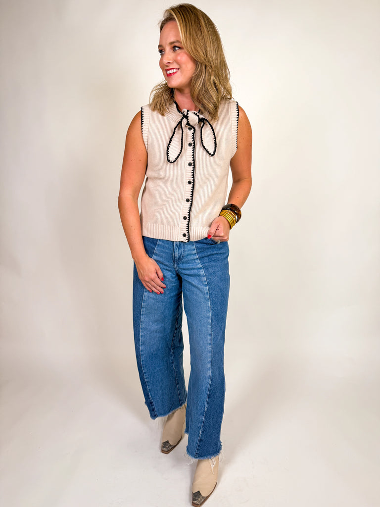 Woman wearing a cream ribbed knit vest with black contrast trim, button front, and bow neckline detail