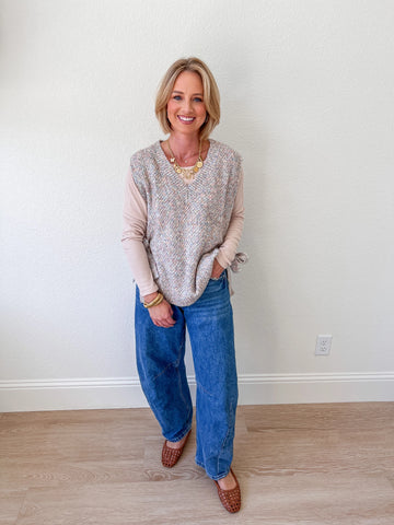 Woman wearing a beige side-tie knit vest over a light pink long-sleeve top, paired with wide-leg denim jeans, standing sideways in a bright indoor setting